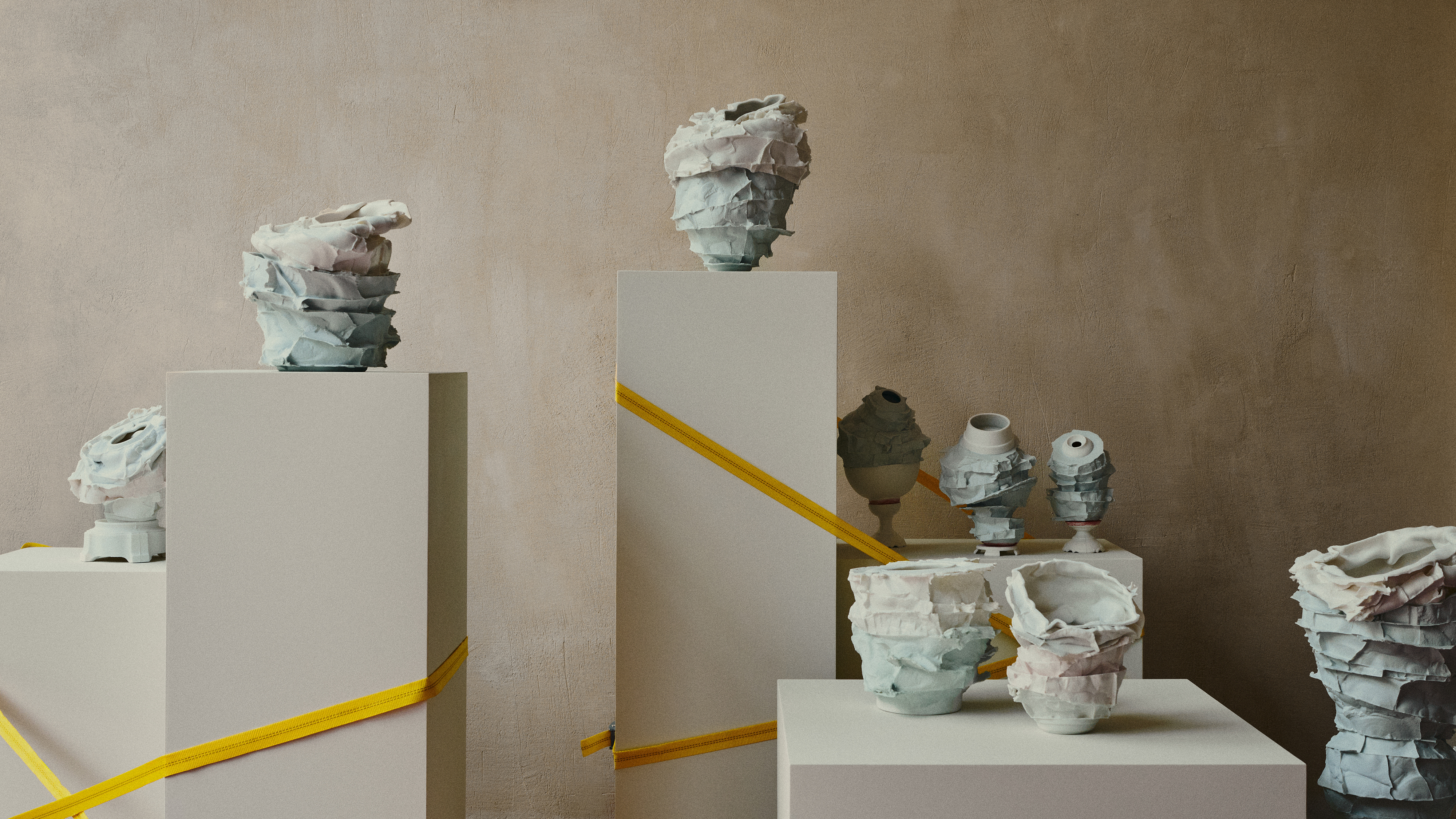 Collection of handcrafted ceramic vases displayed on pedestals with geometric ribbon accents against a neutral-toned wall, creating an artful exhibition.