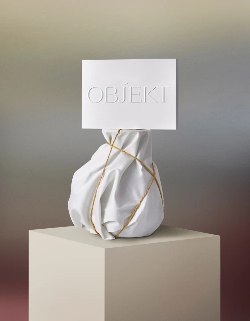 Still life shot of an object wrapped in white cloth and tied with string on a beige pedestal with the word Objekt on a sign.