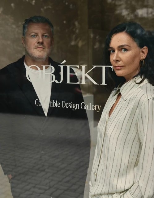 Headshot of a man and woman in front of a glass window with the word 'OBJEKT' on it. The woman has dark hair and is wearing a striped blouse.