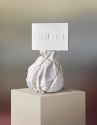 White wrapped object tied with string and topped with debossed paper sign on plinth