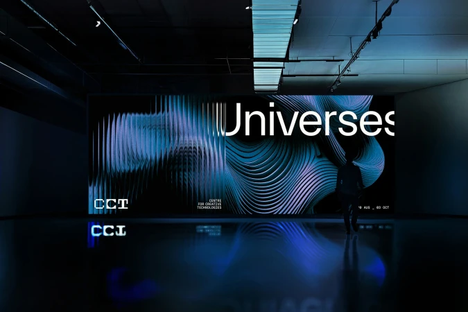 Silhouetted figure stands before a digital art installation featuring vibrant abstract patterns and the word 'Universes' in a dark room.