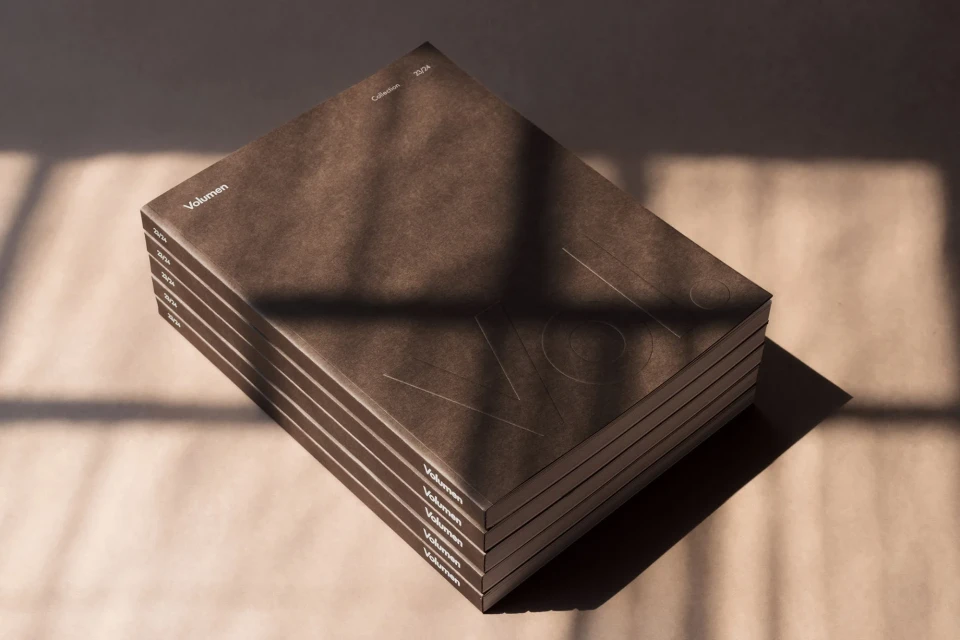Stack of brown Volumen catalogs with subtle design details, casting a shadow on the neutral surface under natural, soft window light.