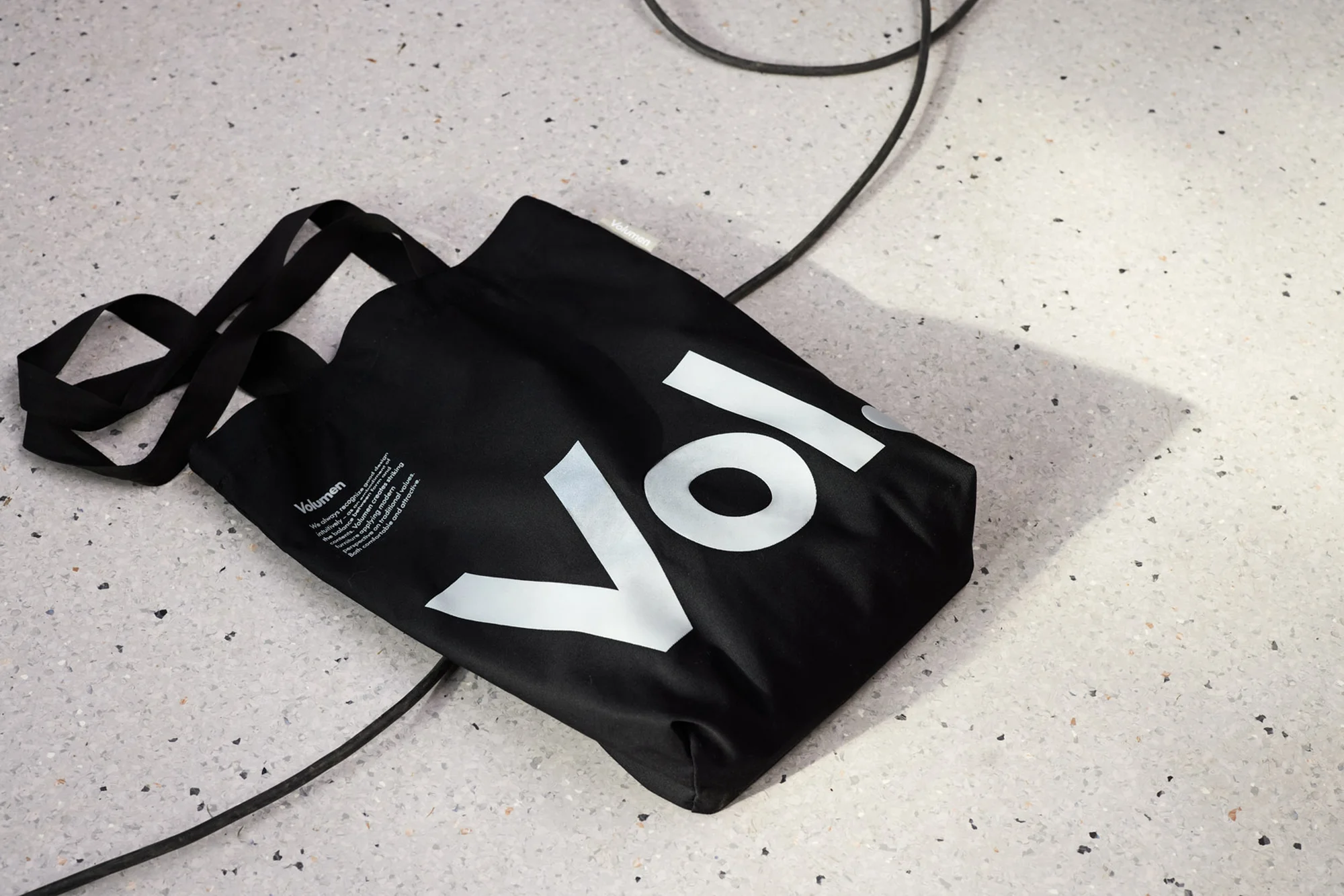 Black tote bag on speckled floor featuring prominent white 'Vol' logo and black electrical cable snaking across speckled flooring.