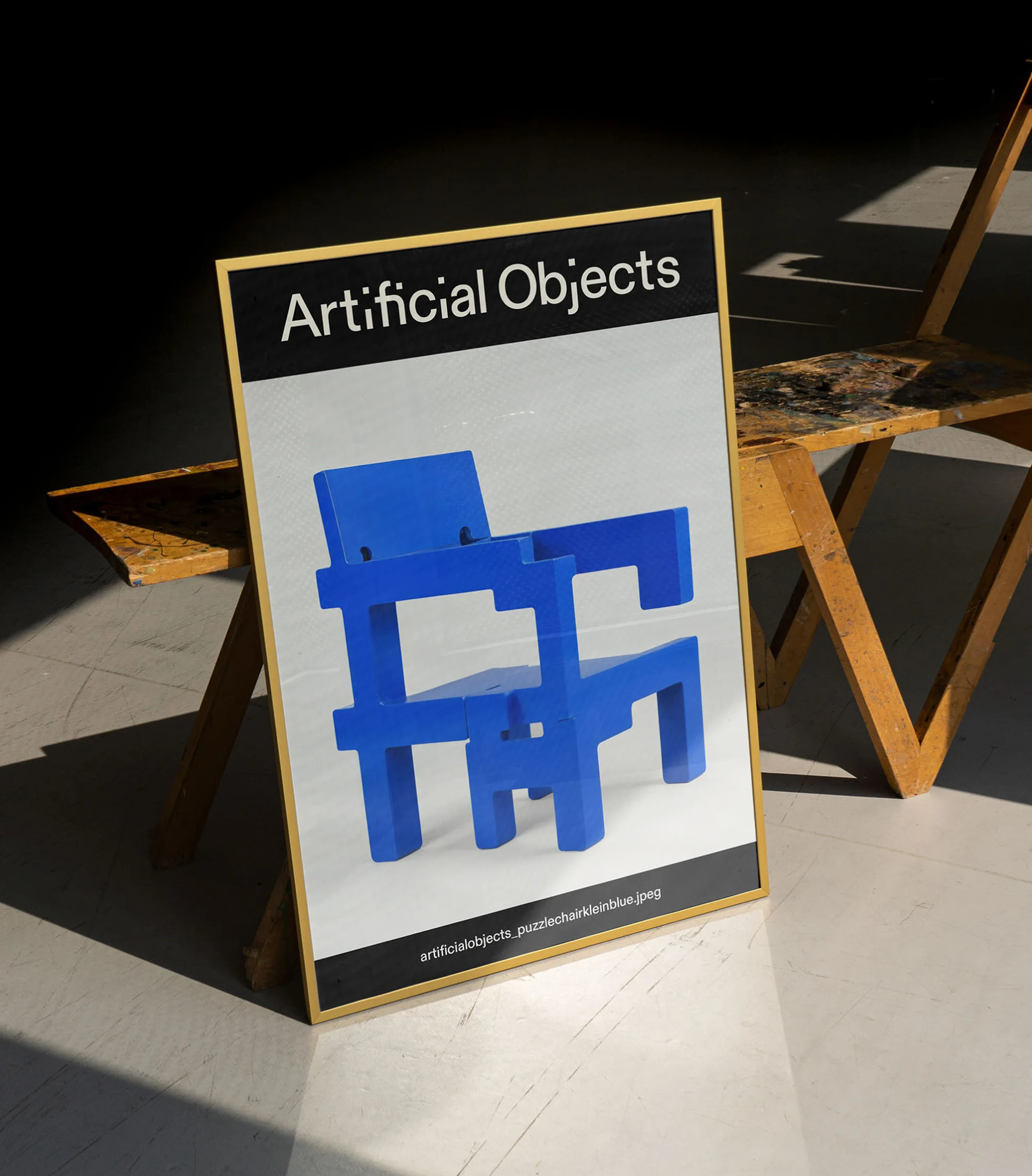 Poster art of a blue geometric chair, displayed indoors against a dark background, adjacent to a light wood-frame easel.