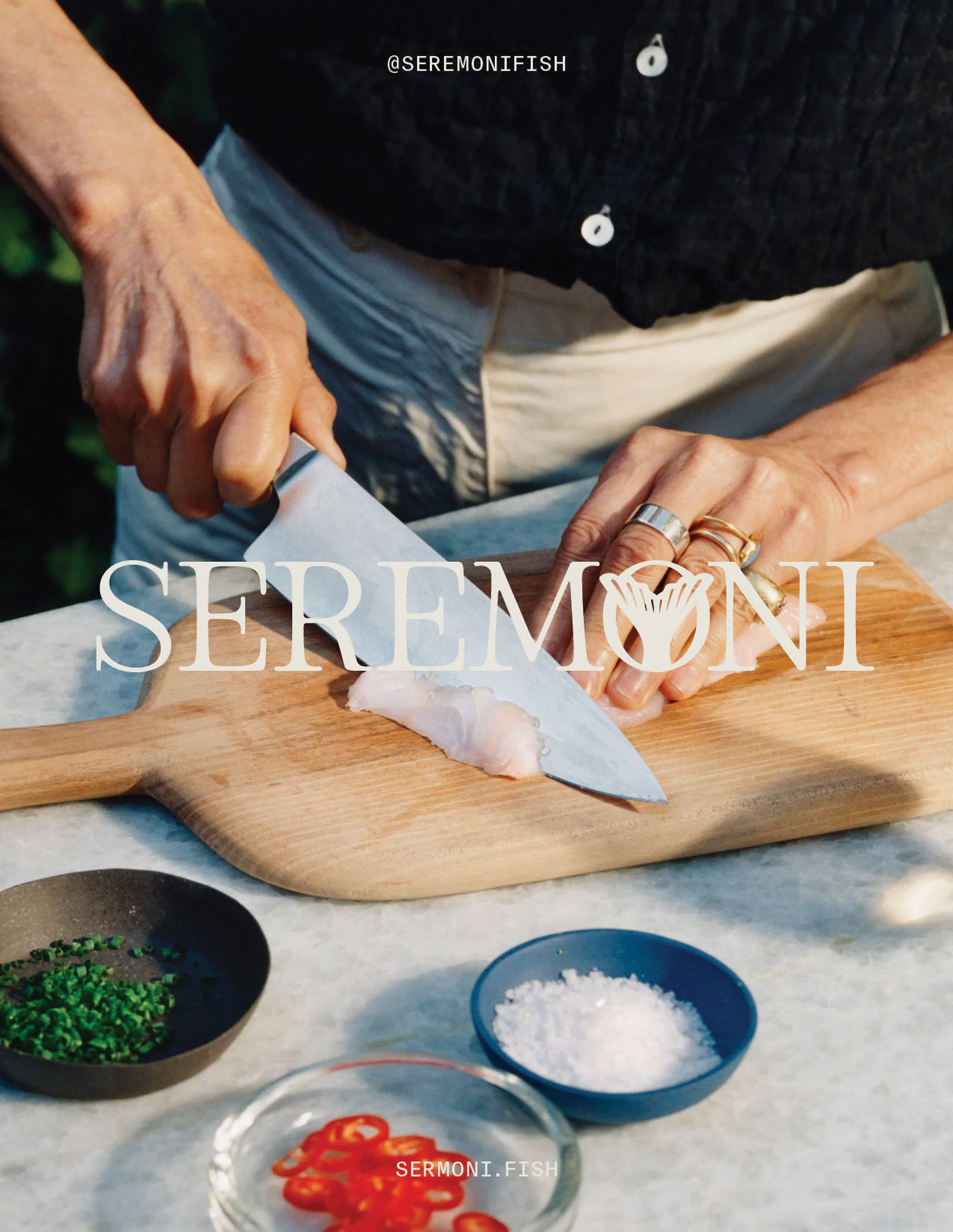 Chef preparing fish on a wooden cutting board with ingredients like chili peppers and herbs, set outdoors with natural lighting.