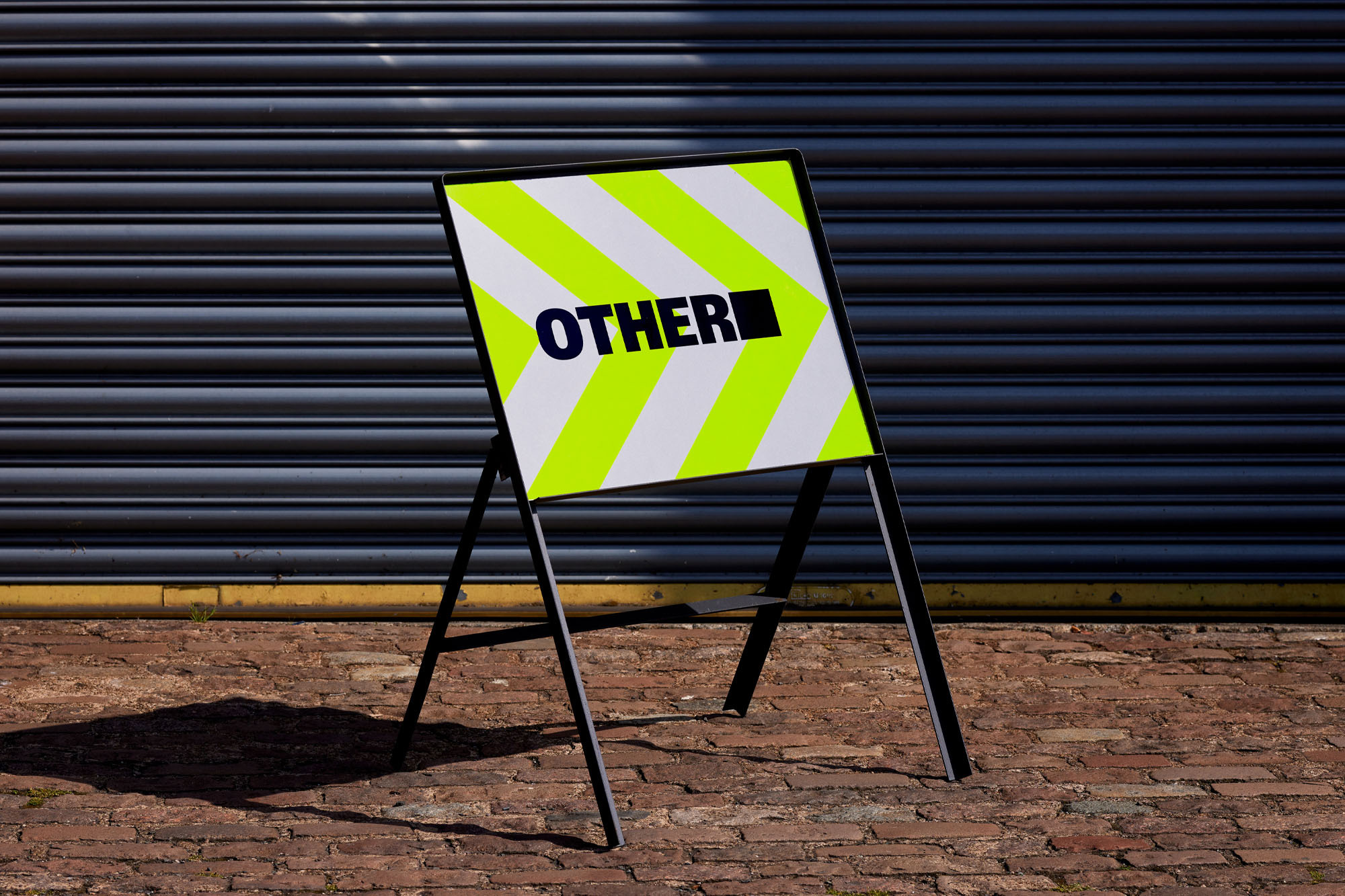 A-frame sign on a brick surface, featuring diagonal neon yellow and white stripes and the word 'OTHER' in bold black letters.