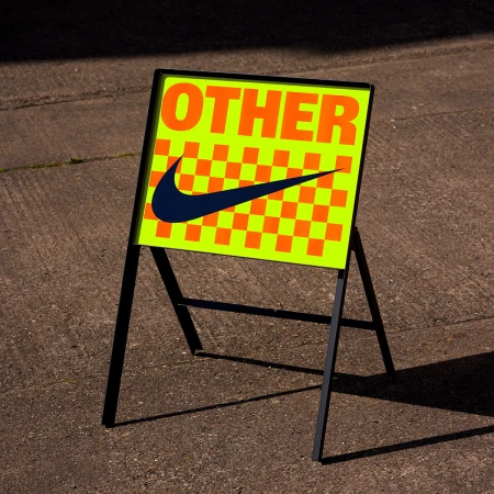 A-frame sidewalk sign featuring bright yellow-green neon backing, orange checkerboard pattern, and bold text reading OTHER with a swoosh logo.