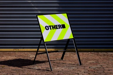 A-frame sign featuring a yellow and white chevron pattern and the text OTHER stands on a cobblestone pavement.