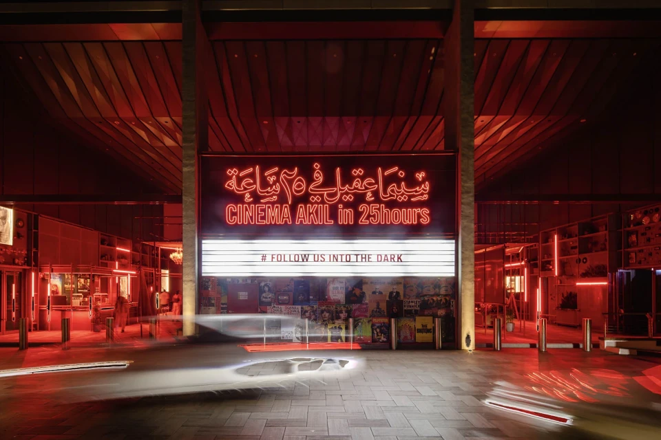 Exterior of Cinema Akil at night, featuring neon signage in English and Arabic, and film posters on a brick wall, brightly lit.