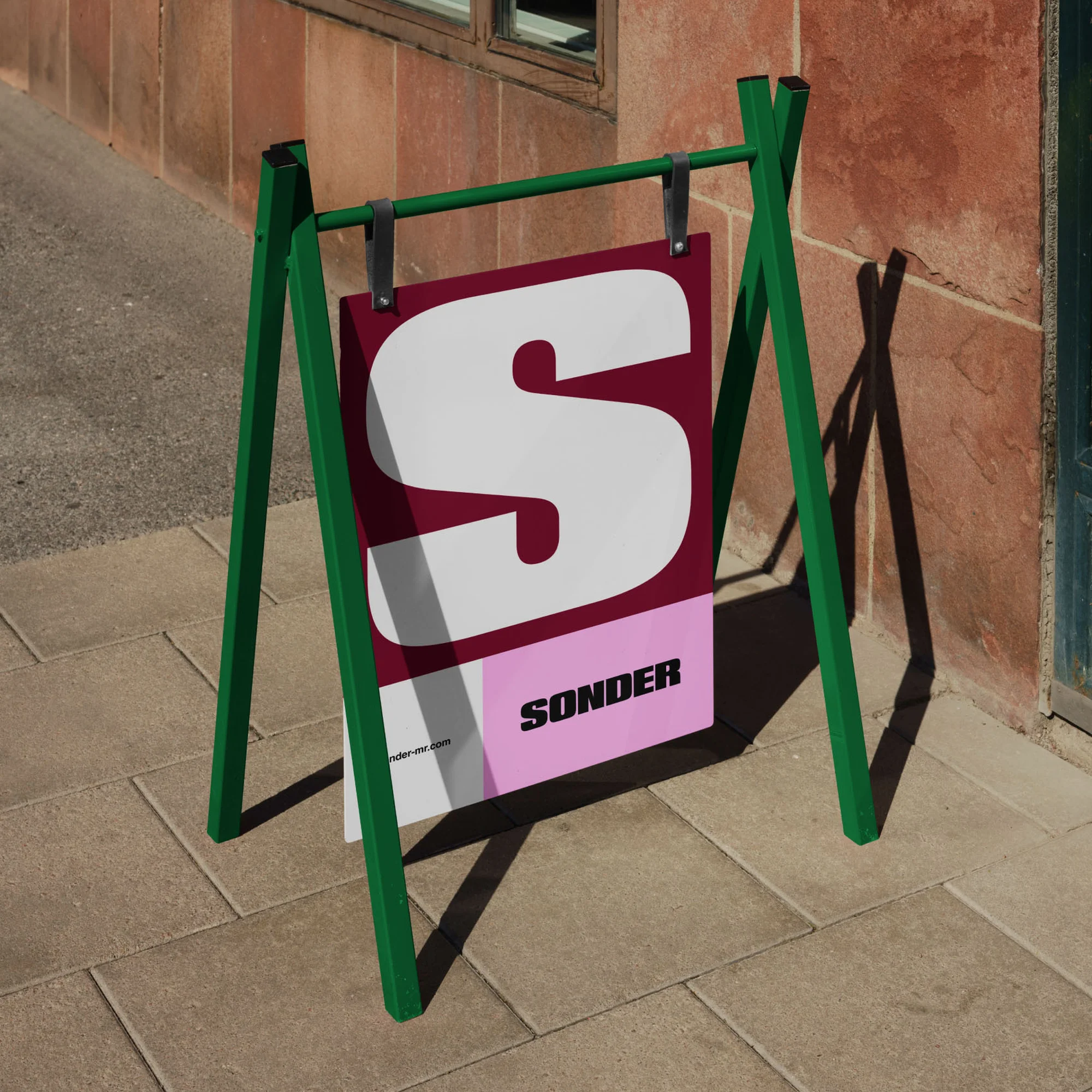 A-frame advertising sign on a sidewalk with the letter S and the word 'SONDER' in bold letters, placed outside against a brick wall.