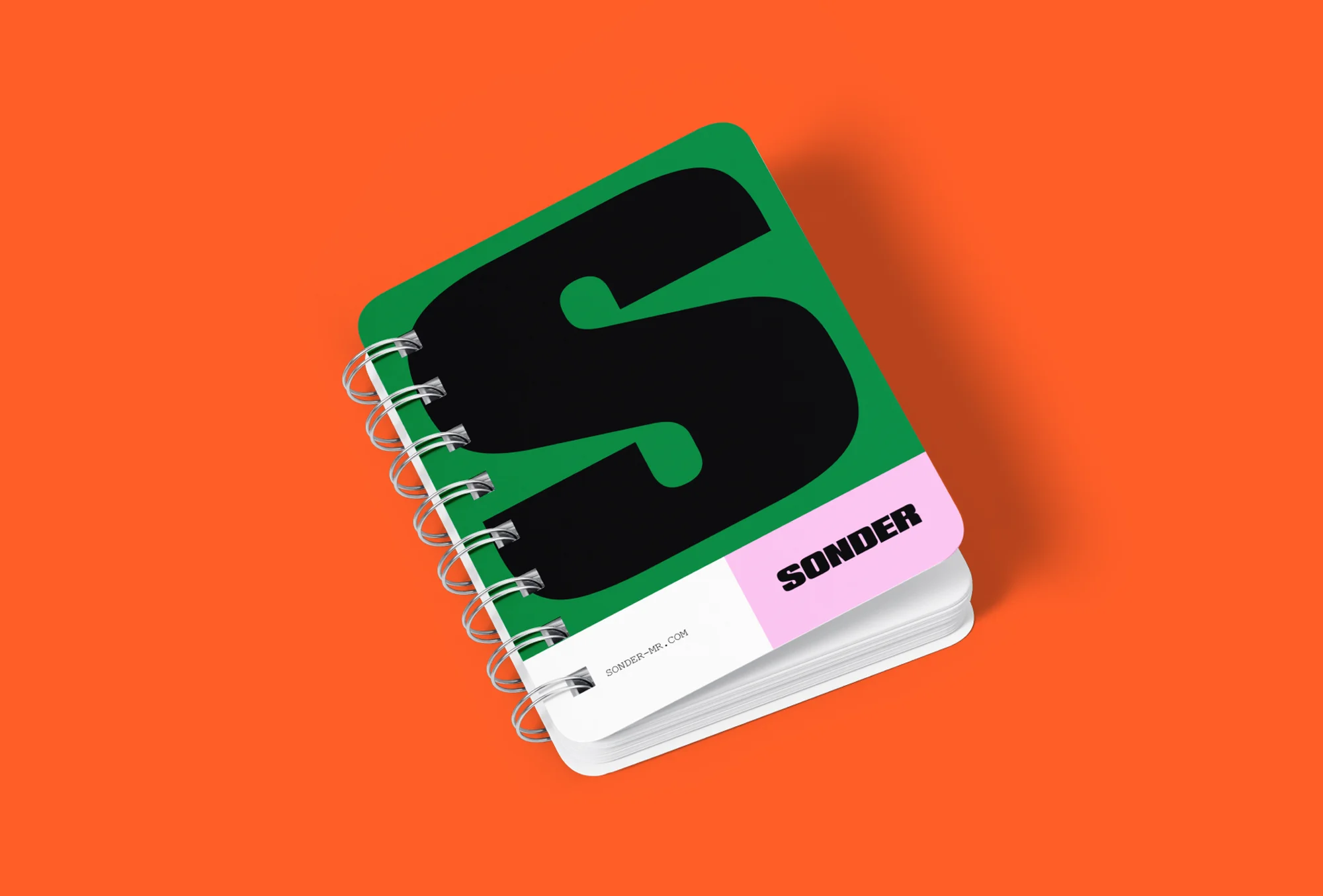 Eye-level, medium shot of a Sonder brand notebook with a green cover, a black 'S' graphic, and a metal spiral binding, set against an orange backdrop.