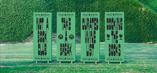 Four green rectangular signs with black typography displayed on a lawn in front of a dense green hedge.