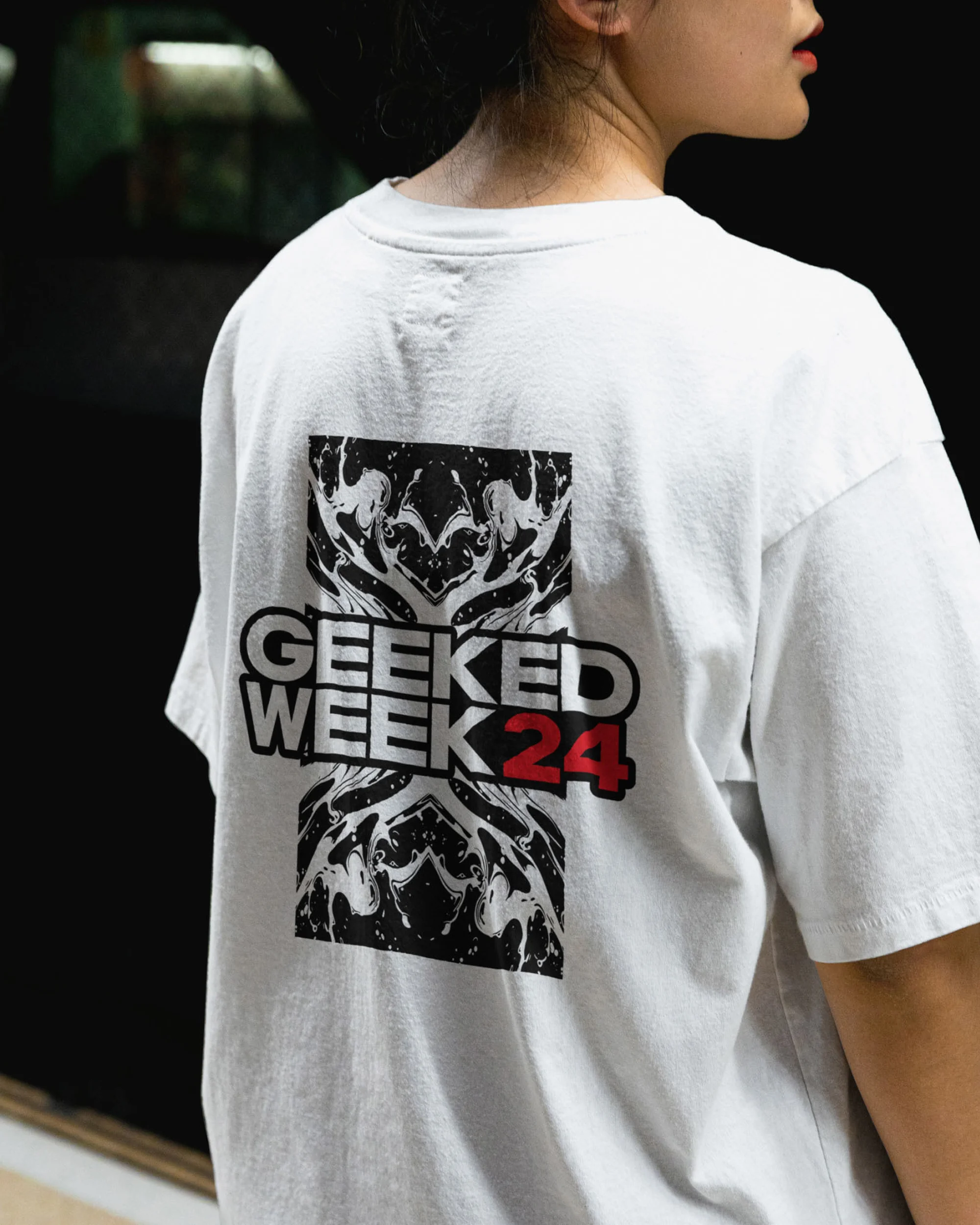 Back of person wearing white t-shirt with graphic design featuring the words 'Geeked Week 24' in black and red against a black background.