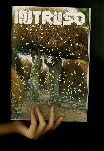 Magazine cover features two people in protective suits carrying a bundle of tumbleweeds, overlaid with an intricate pattern of small circles.