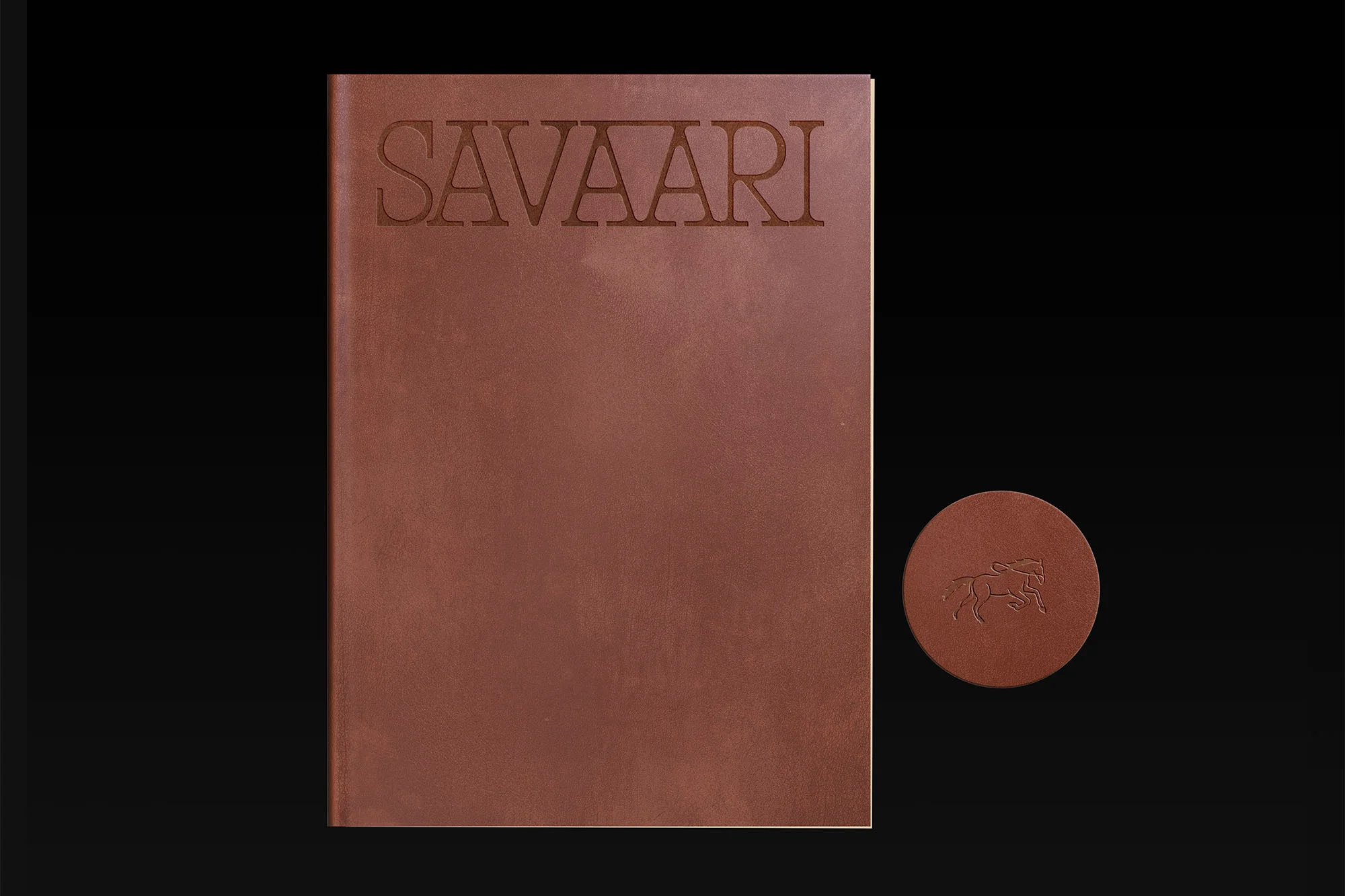 Leather-bound book titled 'SAVAARI' with an embossed horse logo and a matching coaster against a dark background.