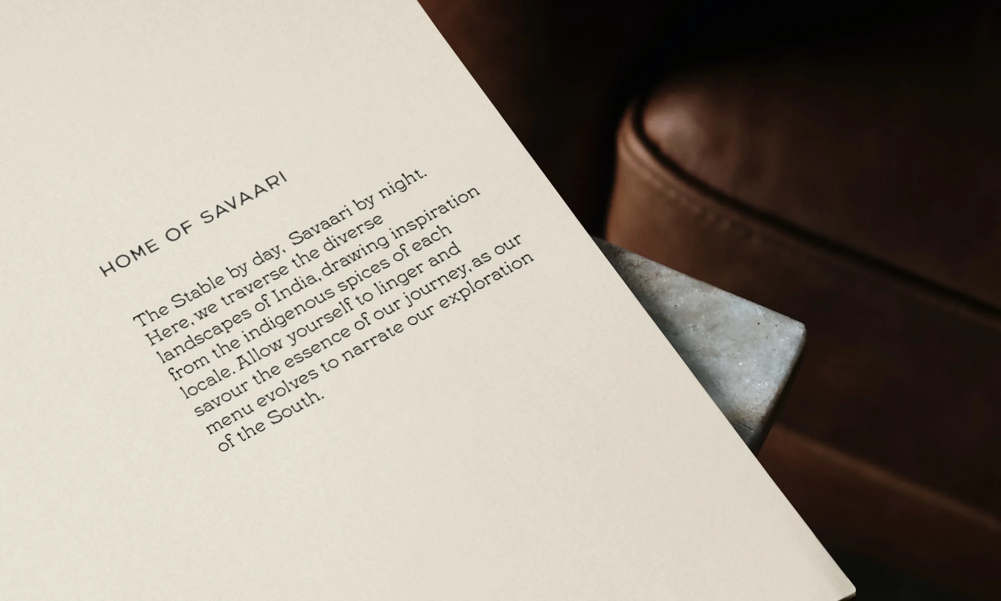 Menu on a table with text 'HOME OF SAVAARI' at an Indian restaurant. Close-up of the paper with a brown chair in the background.