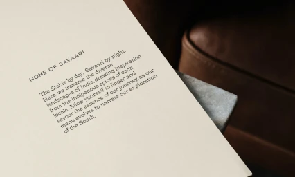 Close-up of a cream-colored menu card for Savaari featuring branded text resting on a concrete surface next to a brown leather seat.