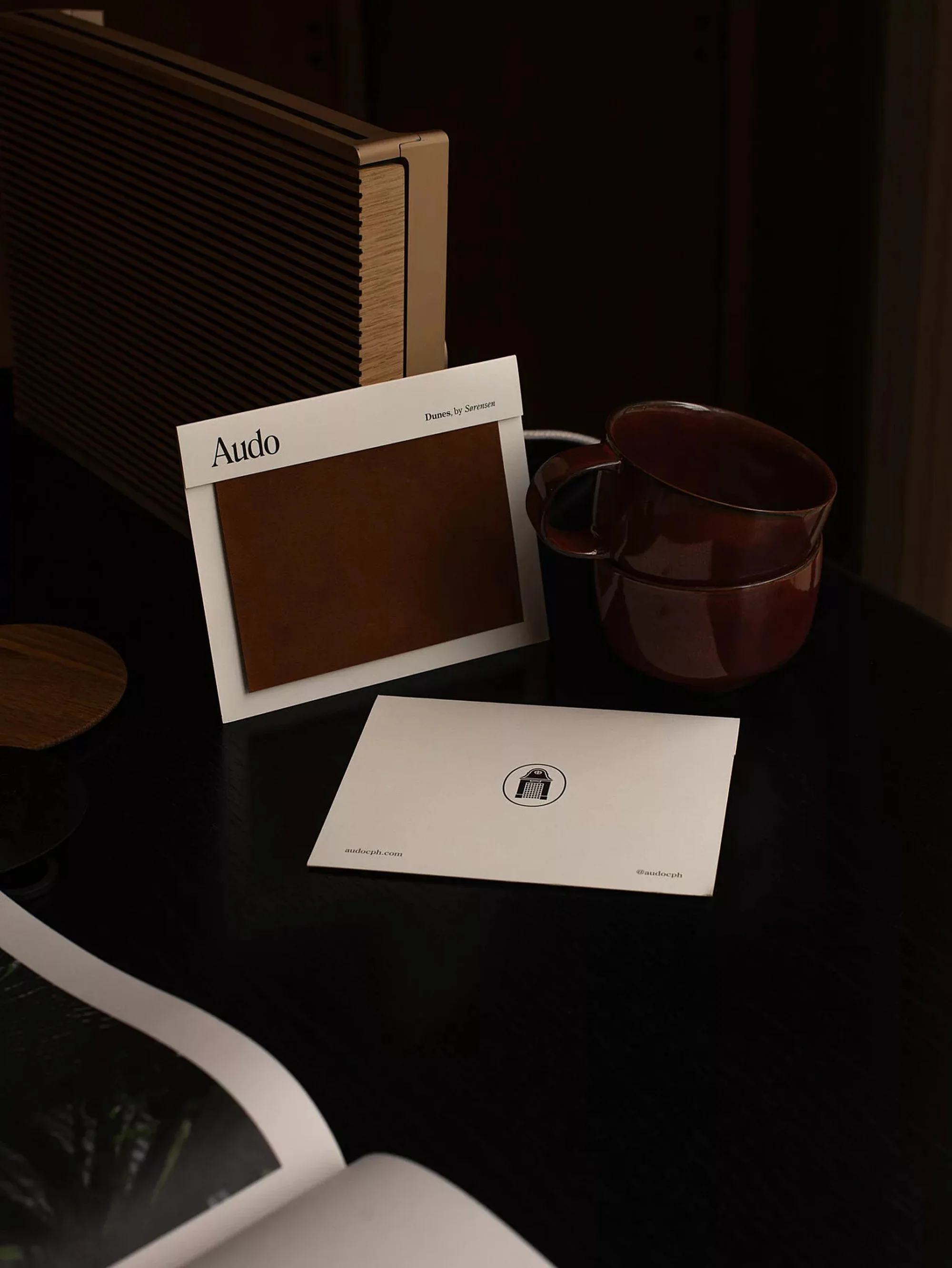 Still life shot of a brown cups stacked next to a color swatch, a coaster, and a book on a black table with soft, diffused lighting.