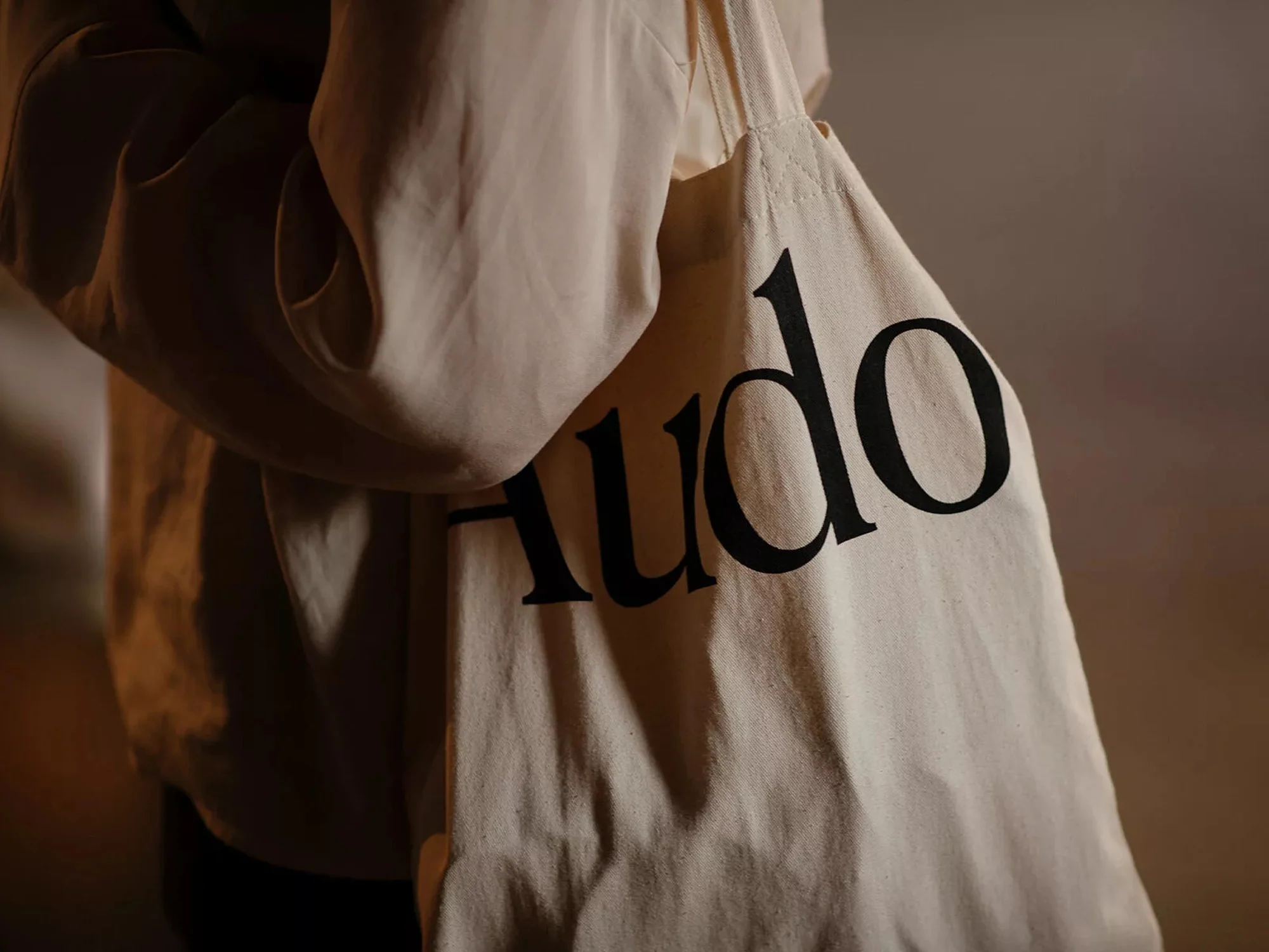 Close-up of a person wearing a tan sweatshirt and carrying a beige tote bag with the word "Audo" printed in black lettering.