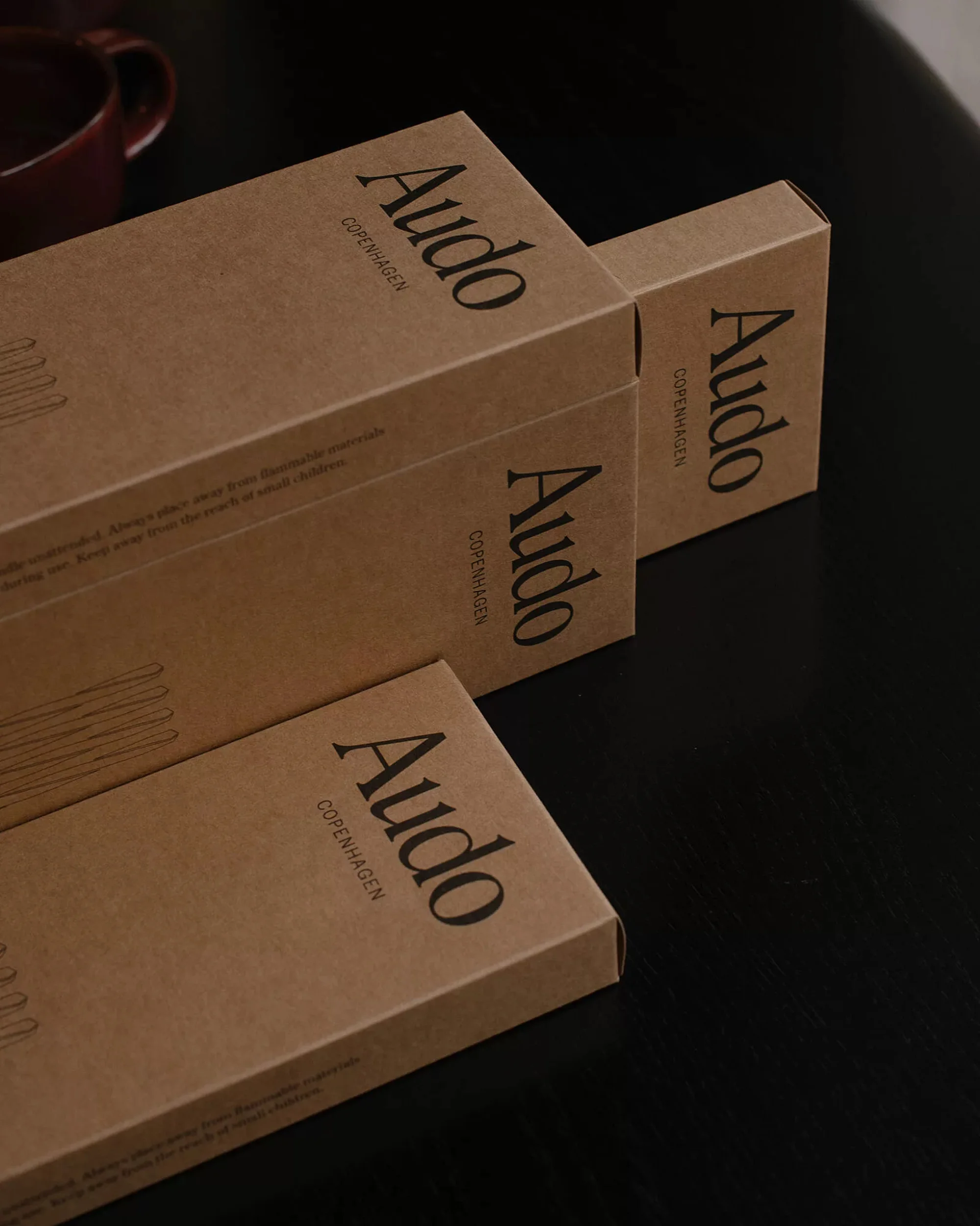 Three brown product boxes from Audo Copenhagen are stacked on a dark surface, with a partial burgundy mug in the background.