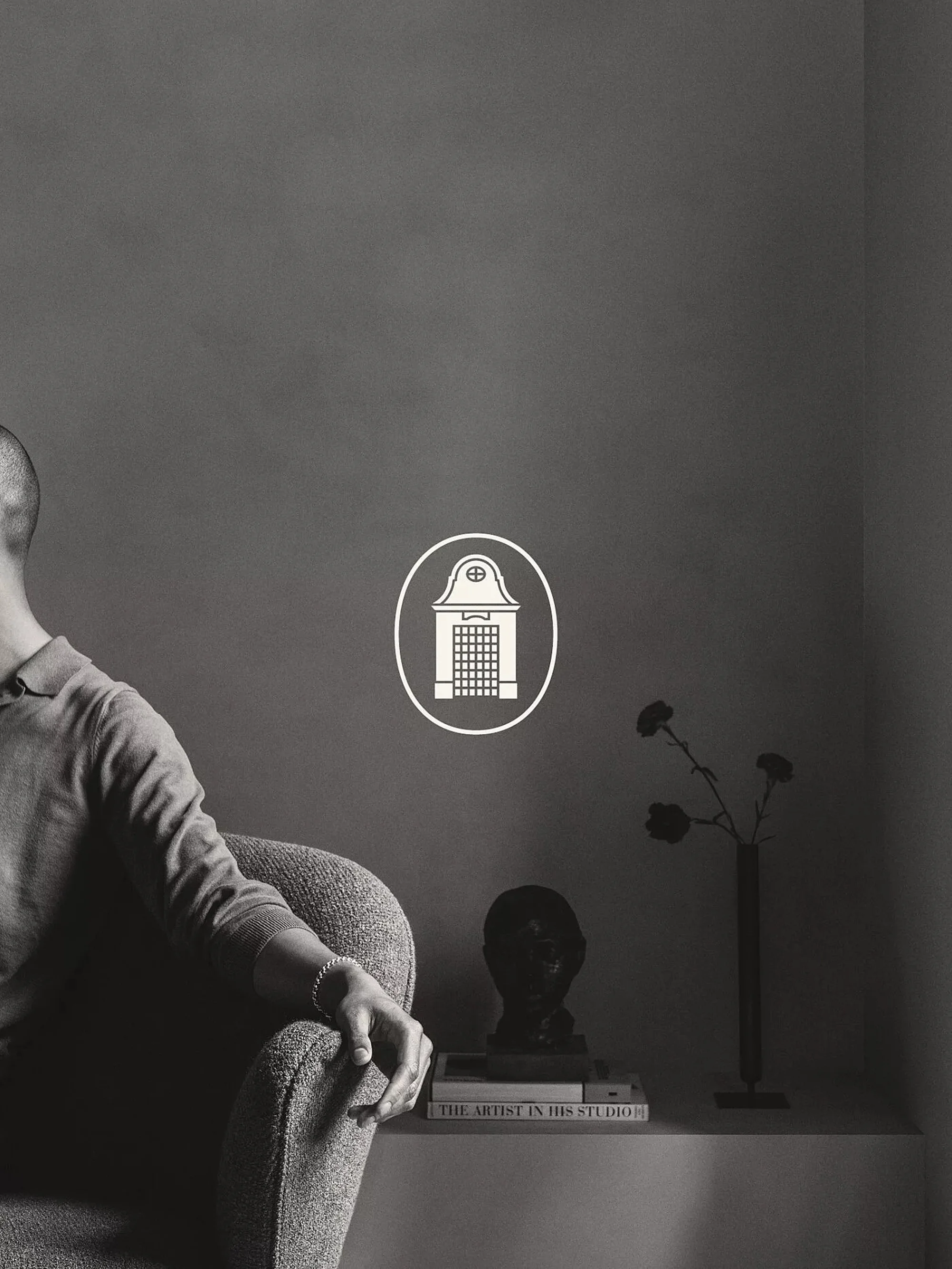 Monochrome image of a person seated in an armchair with a minimalist shelf displaying books, a bust, and a floral vase.