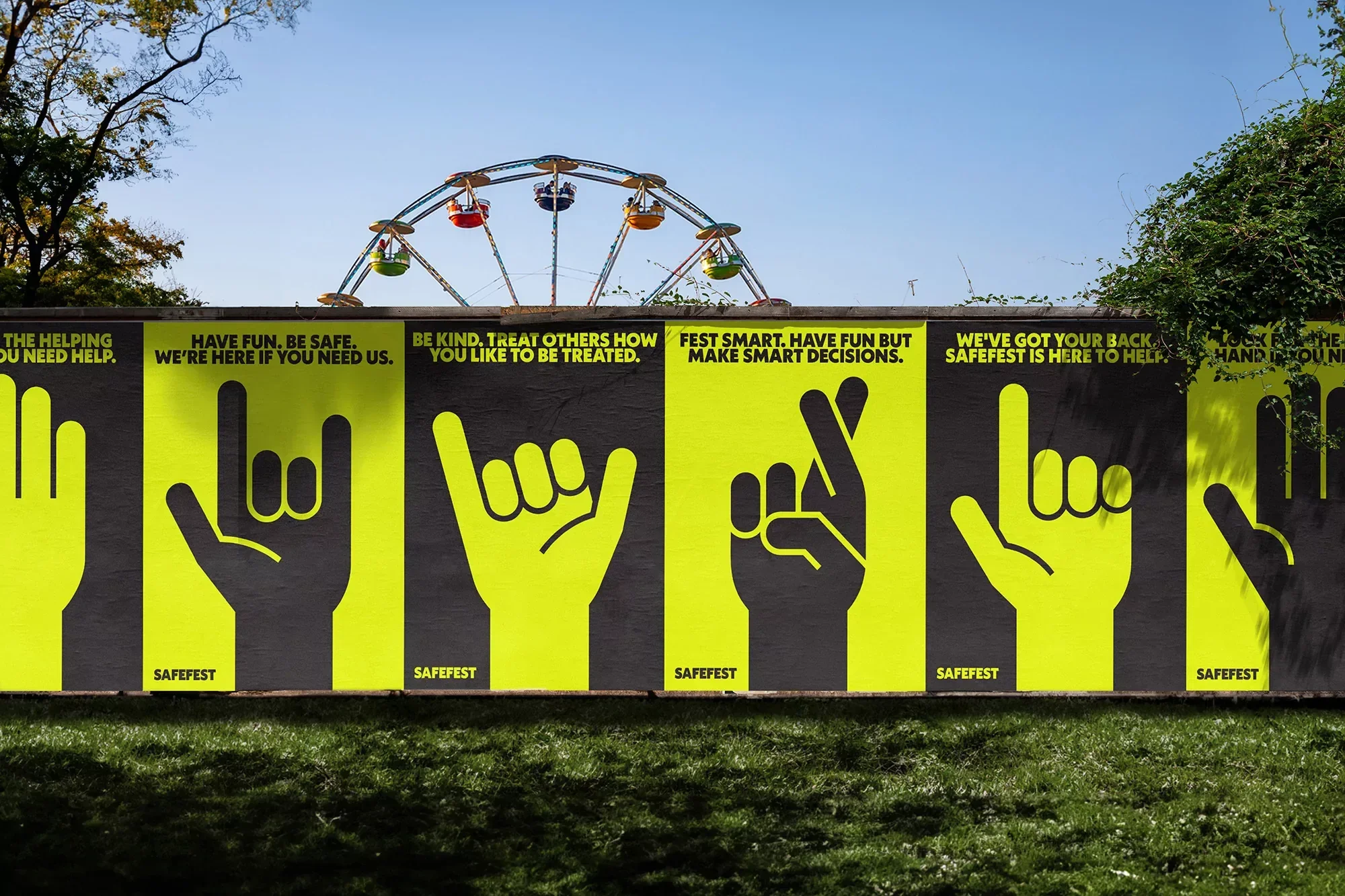 Eye-level medium shot of SafeFest signage on wall with hand gestures and positive messages, Ferris wheel in background, sunny day.