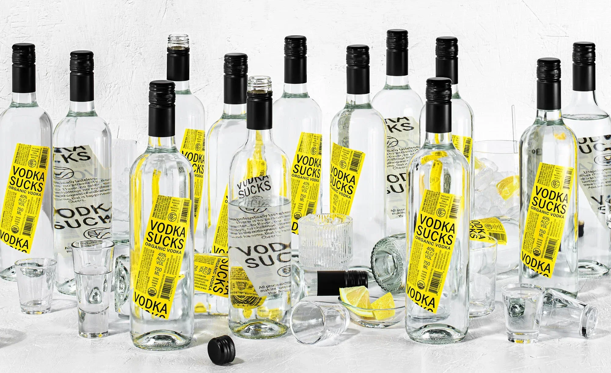 Multiple bottles of Vodka Sucks organic vodka with shot glasses and lemons on a white surface for a product shot.