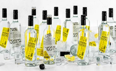 Group of clear glass vodka bottles with bright yellow product labels arranged tightly on a white textured surface.