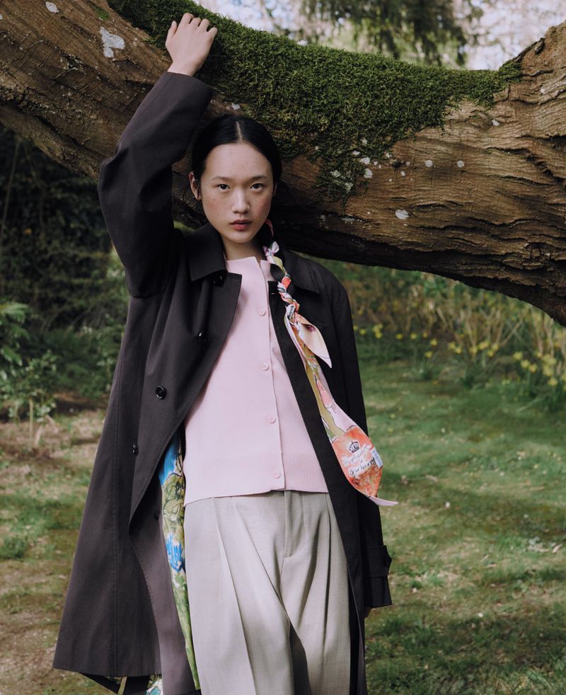 Woman in trench coat posing by mossy tree trunk in outdoor fashion shot. Model wears pink top, khaki pants, scarf braid in the woods.