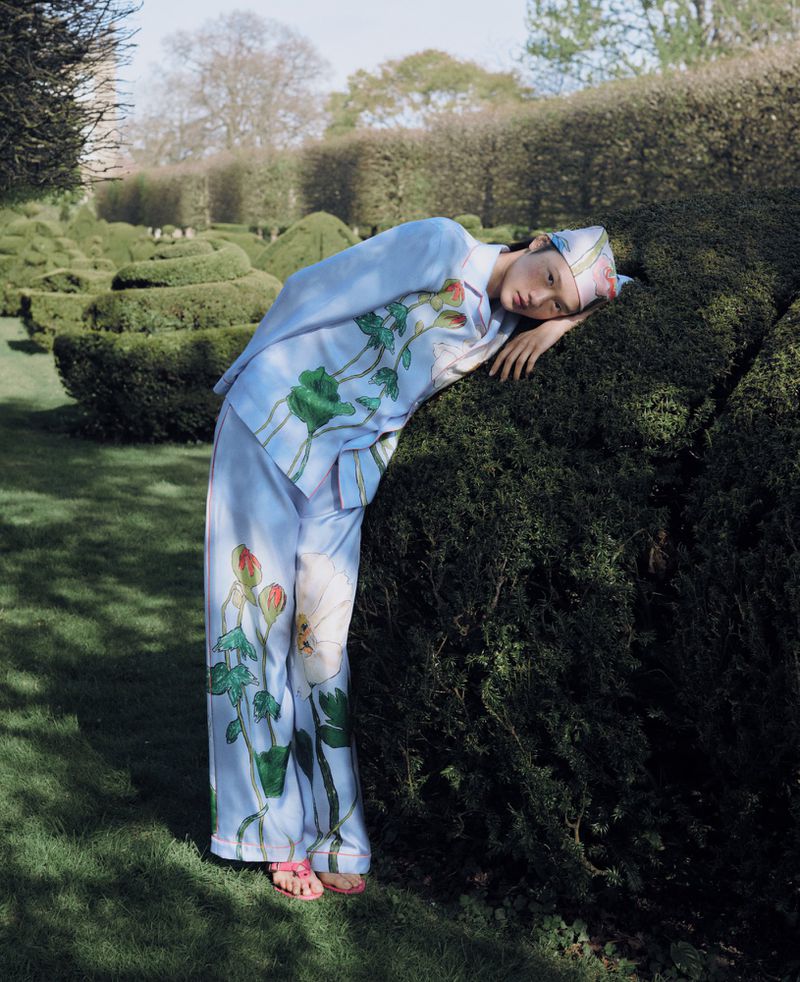 Woman in floral pajamas leans against topiary in an outdoor garden setting with a patterned hedge maze in the background, bathed in sunlight.