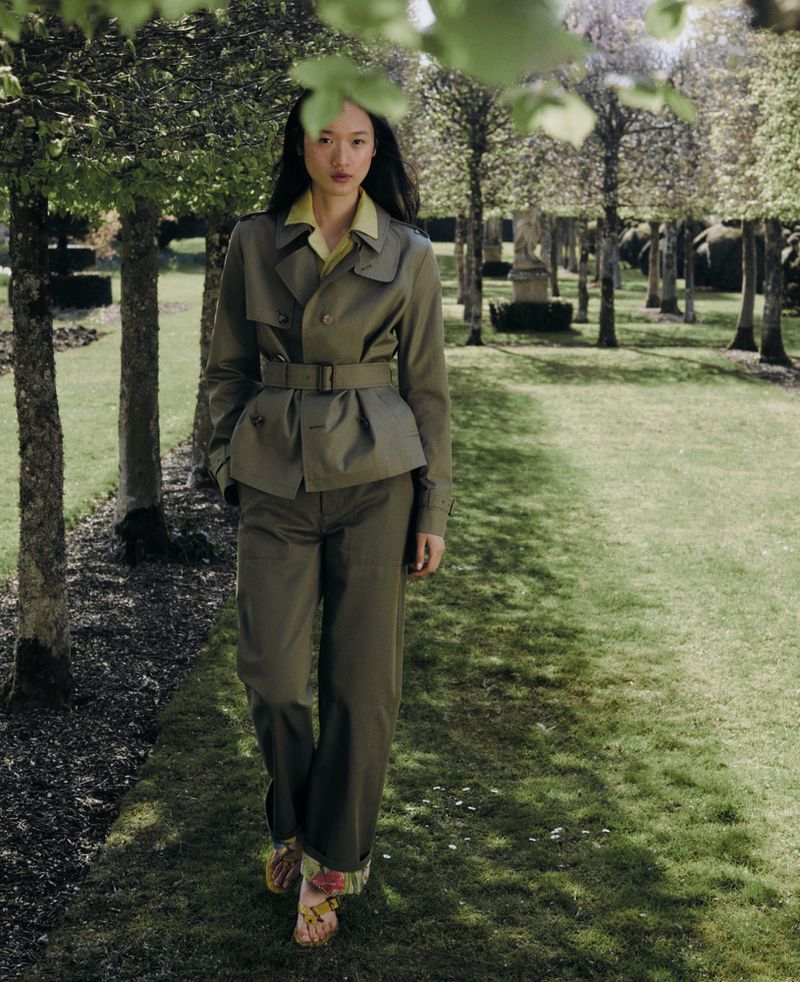 A woman in a belted trench coat and trousers walks on a grassy path in a park with trees, wearing heeled sandals with a floral print.