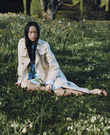 Asian woman sits in grassy field wearing a trench coat. Medium shot features spring flowers, natural light, and an outdoor setting.