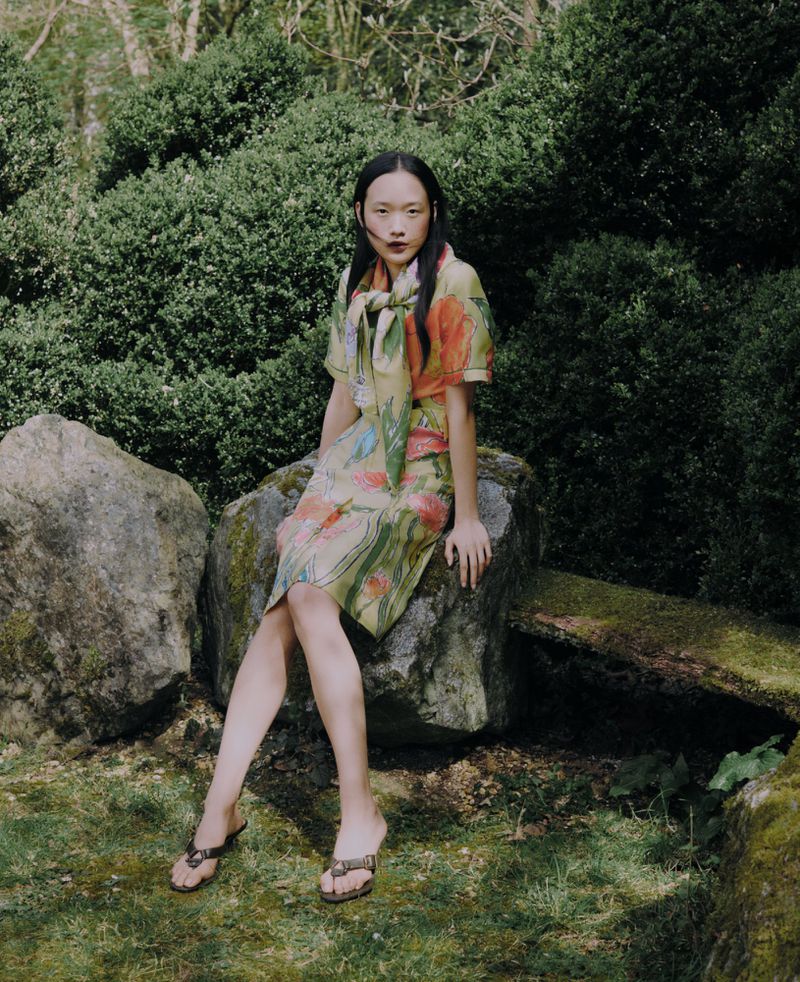 An Asian woman in a floral dress sits on a rock in a garden. She is posing and wears sandals and a floral scarf.