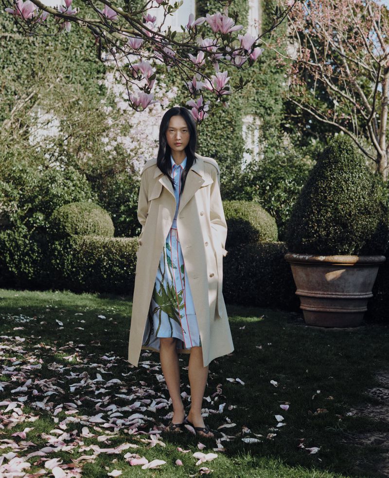 Woman in trench coat stands on grass under magnolia tree. She wears a printed dress and dark shoes. House and garden are in background.