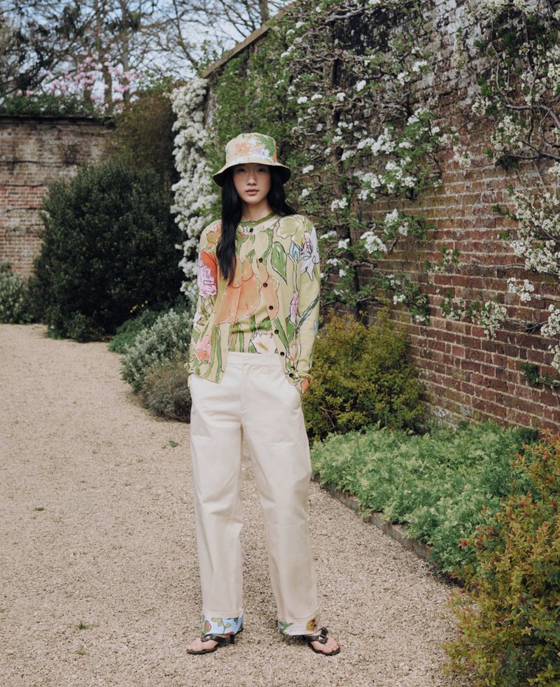 Woman in floral print cardigan and bucket hat stands on gravel path surrounded by green bushes and blossoming brick wall.