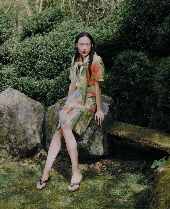 Model Venus He in floral print dress and matching scarf seated outdoors on a mossy garden rock for Burberry X Highgrove