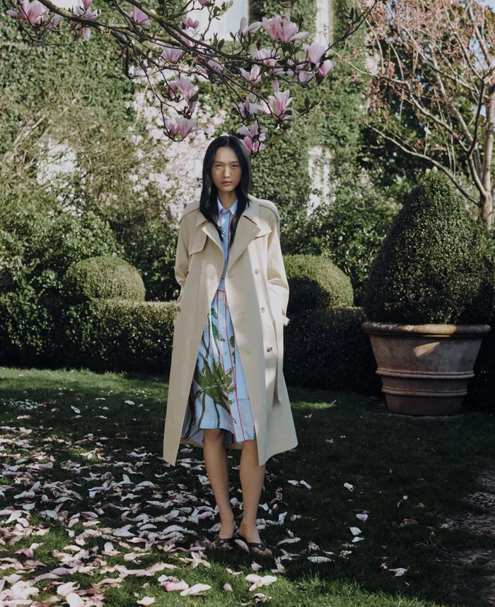 Model standing outdoors wearing a beige trench coat over a patterned dress beneath flowering branches and green topiary hedges