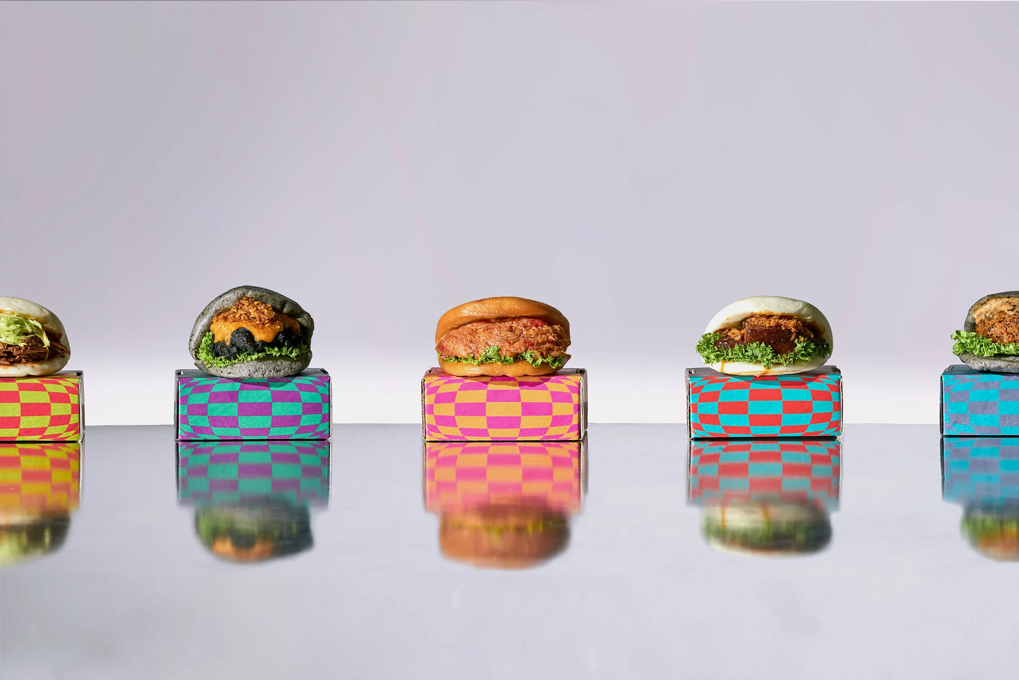 Five different burger slider variations on colorful patterned boxes, evenly spaced on a reflective surface against a neutral background.