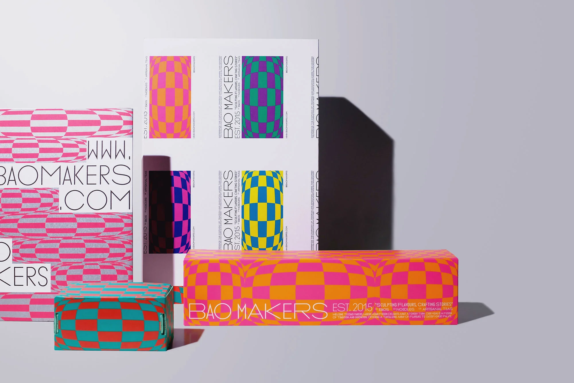 Promotional arrangement featuring Bao Makers branded packaging with a playful, checkerboard pattern in bold colors, on a light gray surface.