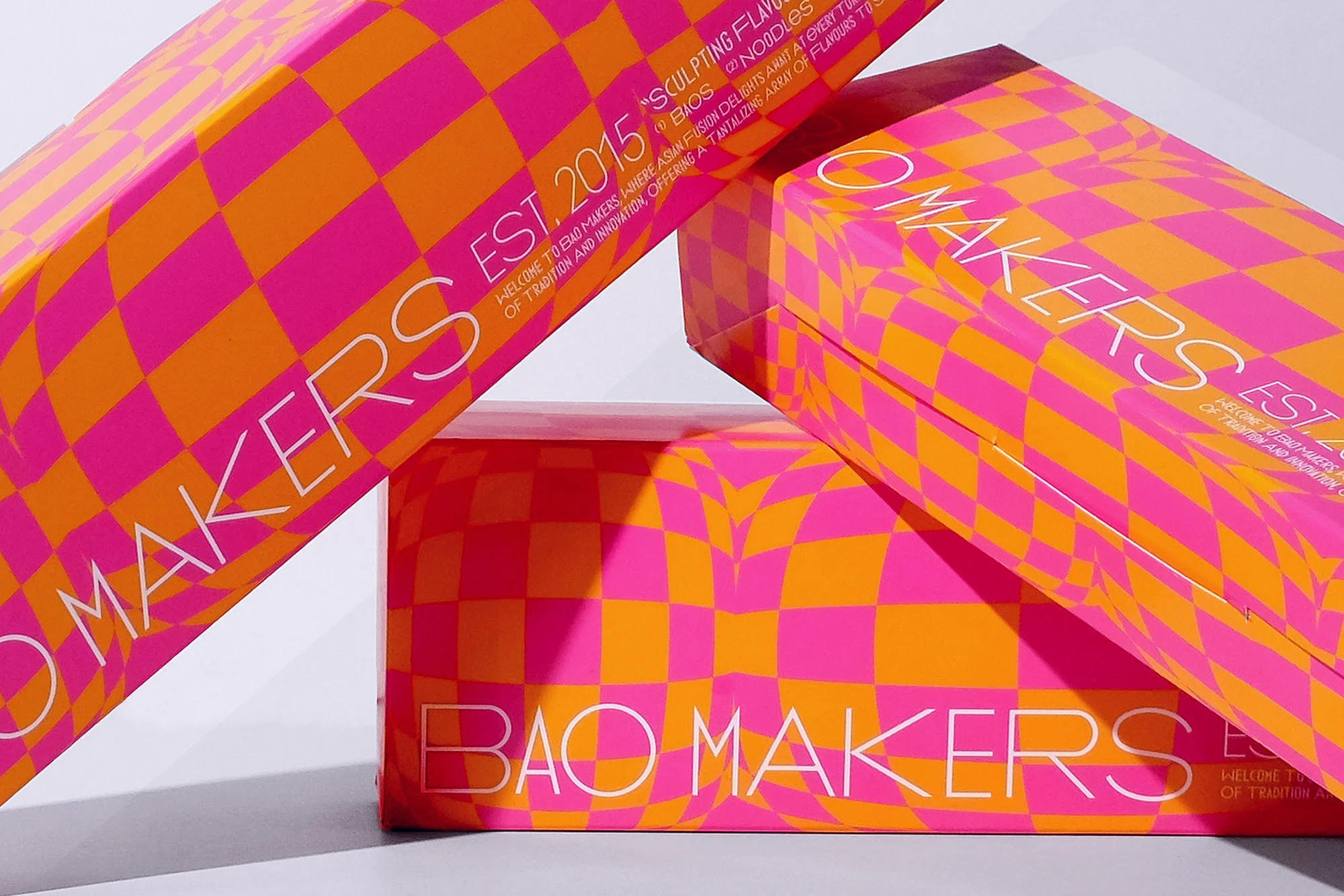 Stacked product boxes with a checkerboard pattern of orange and pink, featuring brand text and establishment year against a white backdrop.