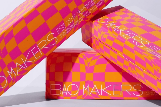 Stacked product boxes with a checkerboard pattern of orange and pink, featuring brand text and establishment year against a white backdrop.