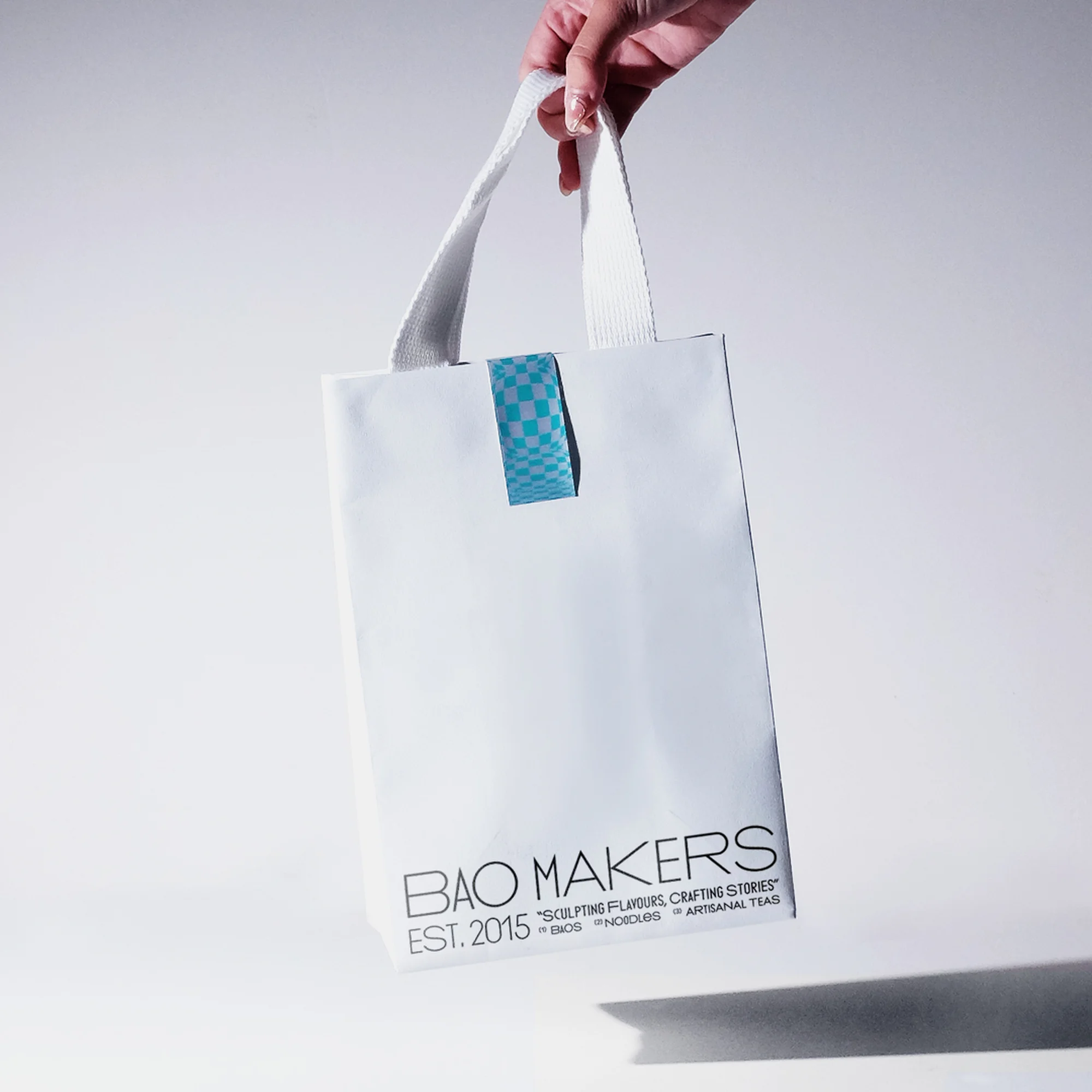 Hand holding a white shopping bag with the text 'BAO MAKERS EST. 2015' on a white backdrop, perfect for food or beverage branding and marketing.