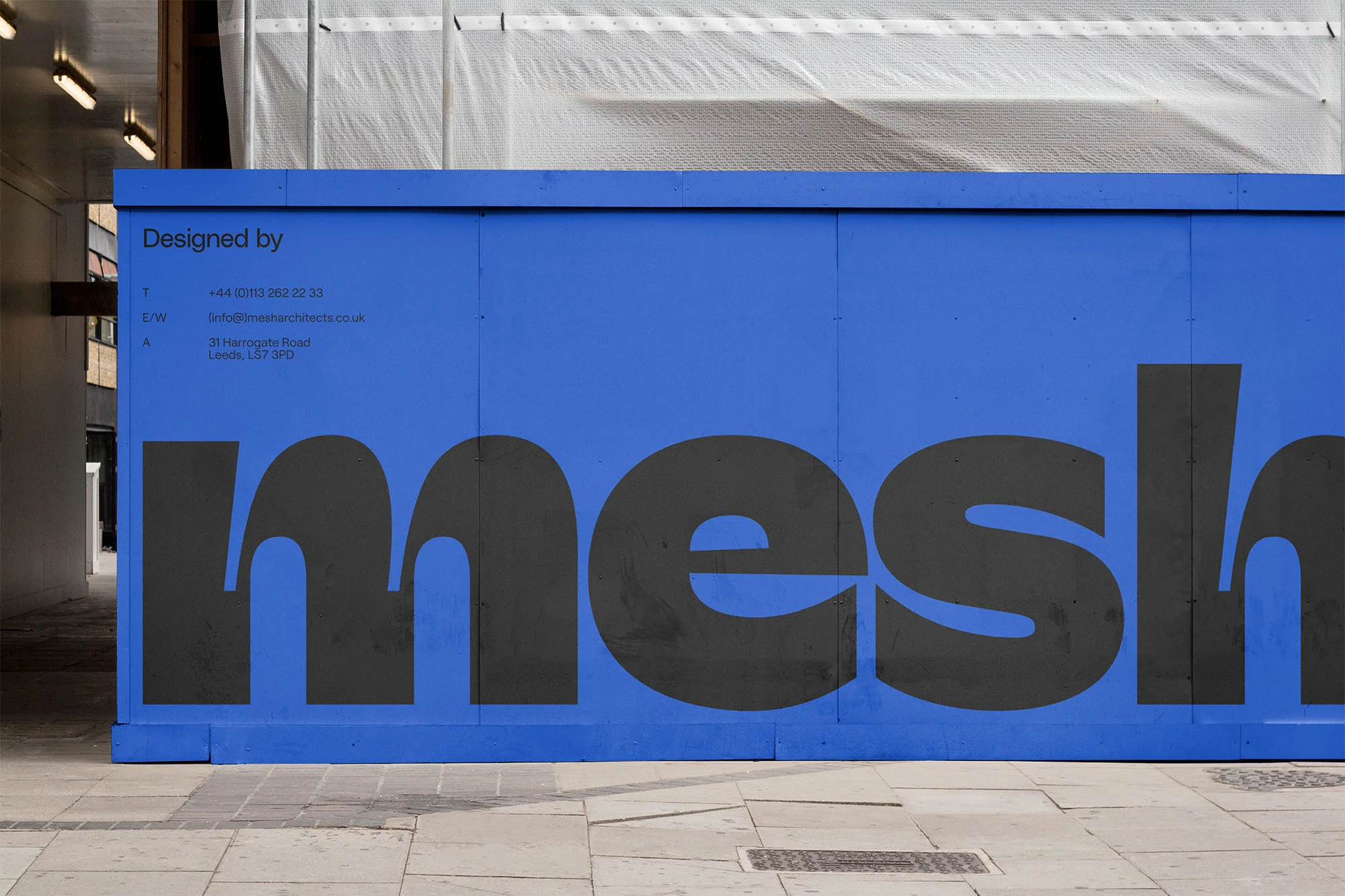 A blue construction hoarding with the name 'mesh' in a large sans-serif font, located on a grey concrete path, photographed in daylight.
