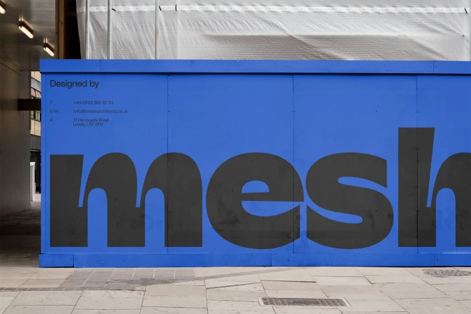 A blue construction hoarding with the name 'mesh' in a large sans-serif font, located on a grey concrete path, photographed in daylight.