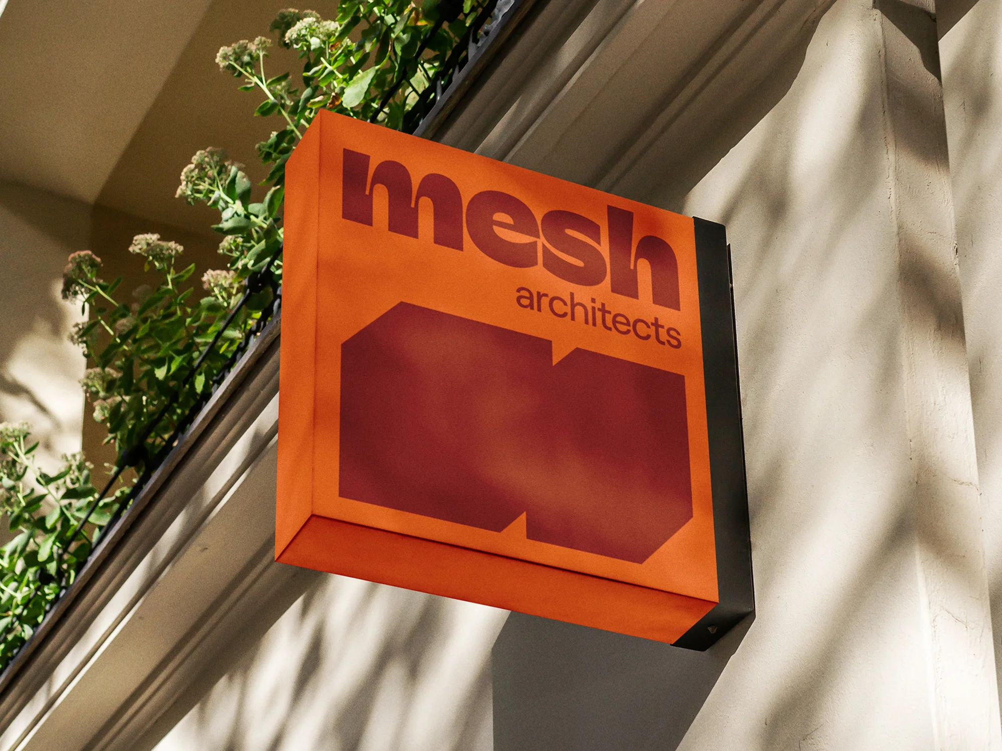 Orange building sign for Mesh Architects, featuring the company logo and name, attached to the side of a light-colored building.