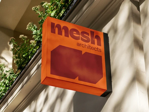 Orange building sign for Mesh Architects, featuring the company logo and name, attached to the side of a light-colored building.