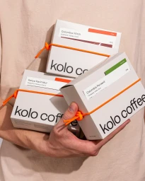 Person holding three white coffee boxes branded with black text, secured with bright orange elastic bands against a neutral background