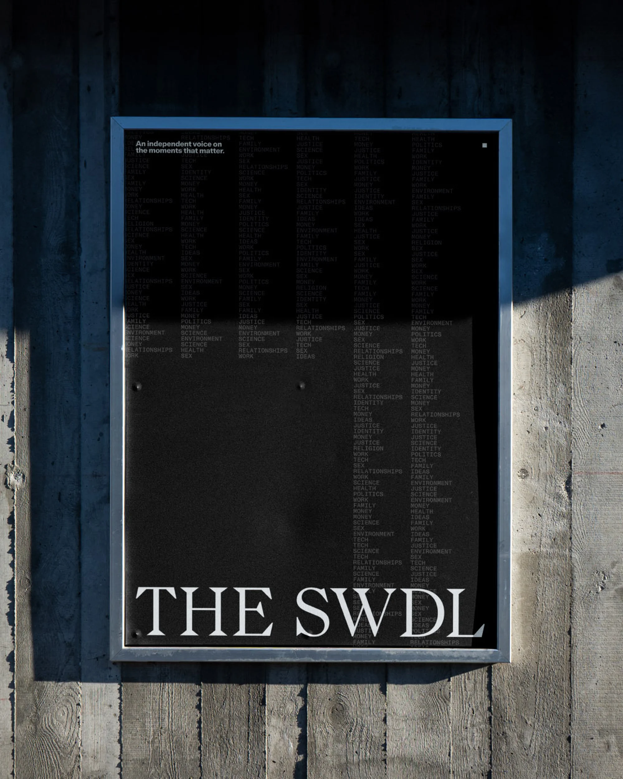 Black and silver metal sign on concrete wall, featuring white text 'The SWDL' and list of additional text, viewed in daytime lighting.