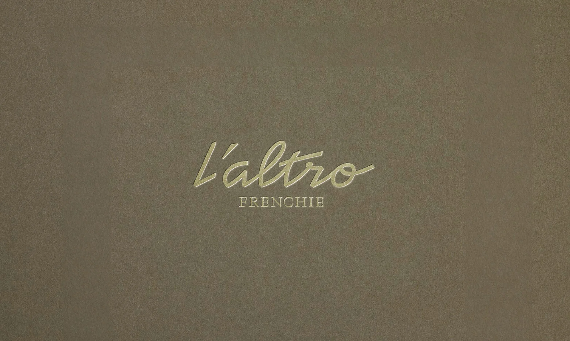 Close-up of gold 'L'altro Frenchie' logo on a neutral grey textured background. Minimalist design with clear typography.
