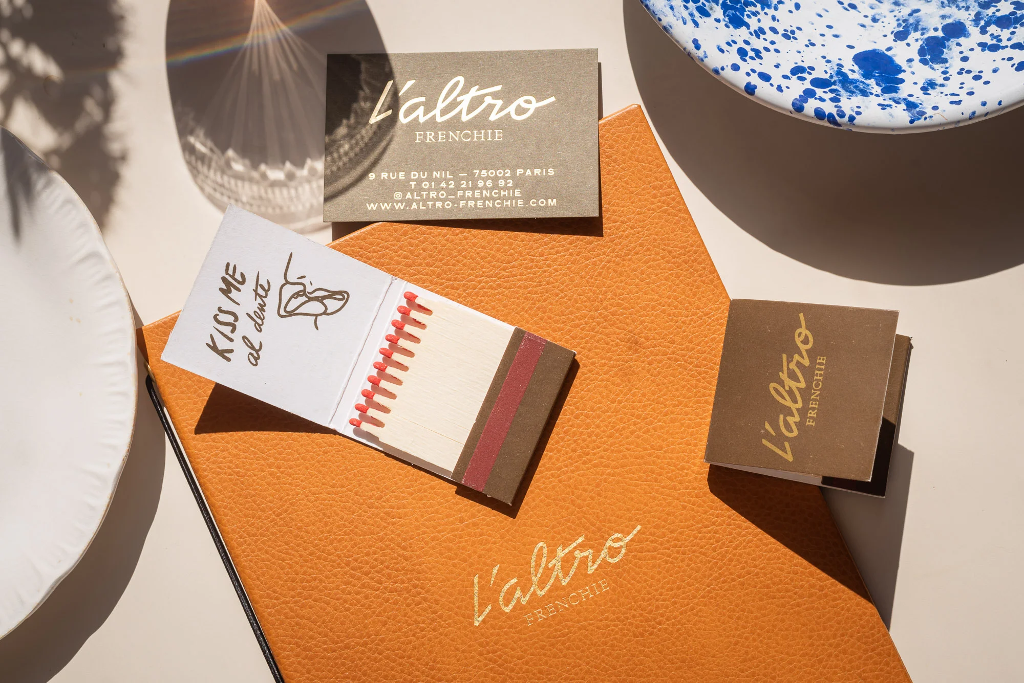 Overhead shot of L'Altro Frenchie restaurant promotional material: menu, business cards, and matchbook on a bright, sunlit table.